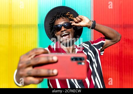 Ottimista giovane tatuato afroamericano maschio in camicia a righe luminose e cappello con occhiali da sole alla moda e orologio da polso che prende selfie su telefono cellulare in piedi contro una parete colorata Foto Stock