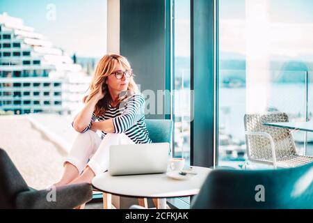 Ritratto di donna matura fiduciosa che si rilassa nella poltrona e sogna mentre si guarda fuori la finestra nella sua casa moderna. Foto Stock