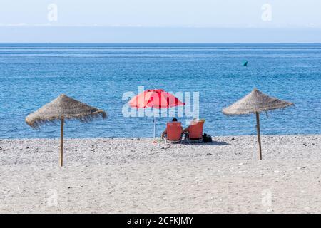 Spiaggia andalusa una soleggiata mattina estiva con due persone a sedere sotto un ombrello rosso che si affaccia sul mare Foto Stock