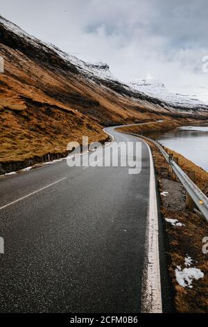 Spettacolare vista della strada tortuosa bagnata situata vicino al fiume e. Pendio di montagna con erba secca in inverno sulle Isole Faroe Foto Stock