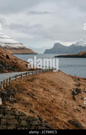 Spettacolare vista della strada tortuosa bagnata situata vicino al fiume e. Pendio di montagna con erba secca in inverno sulle Isole Faroe Foto Stock