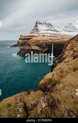 Scenario mozzafiato di case residenziali situate vicino al fiume in altopiano Pendenza scogliera con montagne coperte di neve sulle Isole Faroe Foto Stock