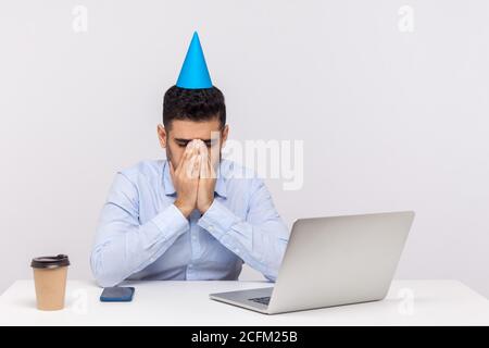 Il ragazzo depresso del dipendente sconvolto con il cono del partito sulla testa che grida sul posto di lavoro, nascondendo faccia che si sente disperato solo il compleanno deve rimanere in ufficio. indo Foto Stock