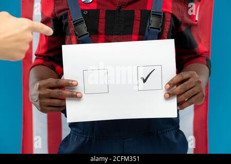 Giovane riparatore nero che tiene la carta di voto con il segno in quadrato Contro la bandiera degli Stati Uniti Foto Stock