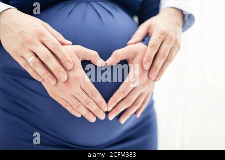Le mani dell'uomo toccano le mani della donna incinta a forma di cuore sul suo bump del bambino. Foto luminosa su sfondo bianco Foto Stock