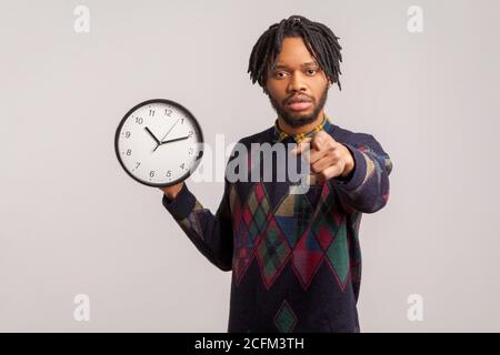 Uomo africano serio bossy con i Dreadlock che tengono l'orologio da parete in mano che punta il dito alla macchina fotografica con lo sguardo concentrato, il tempo all'azione, la motivazione. IND Foto Stock