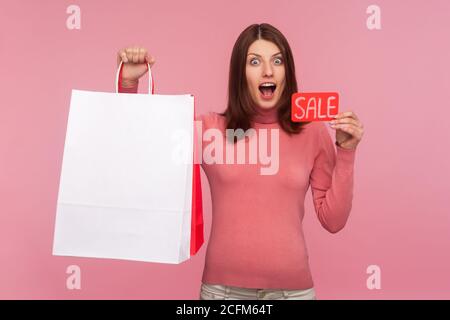 Donna estremamente felice sorprende che tiene nelle mani borse di shopping di carta e carta di vendita, guardando le macchine fotografiche con gli occhi grandi e la bocca aperta. Monolocali interni Foto Stock