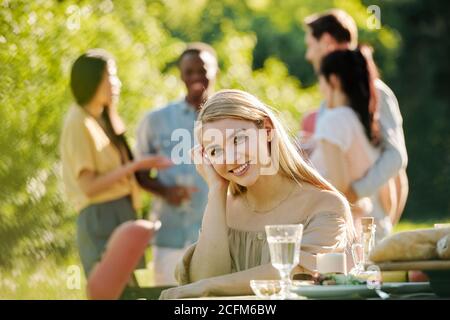 Giovane donna sorridente con lunghi capelli biondi che ti guarda seduto al tavolo Foto Stock