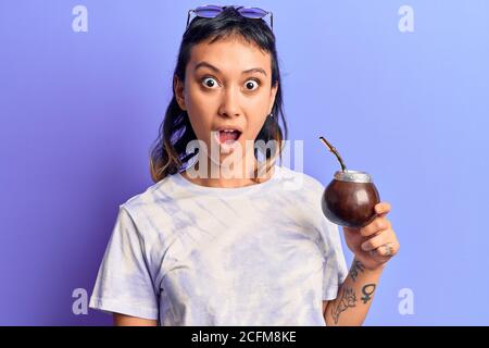 Giovane donna bevendo l'infusione di compagno spaventato e stupito con bocca aperta per sorpresa, faccia incredula Foto Stock