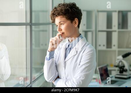Giovane scienziata pensiva femminile in piedi da parete di vetro contro di lei luogo di lavoro Foto Stock