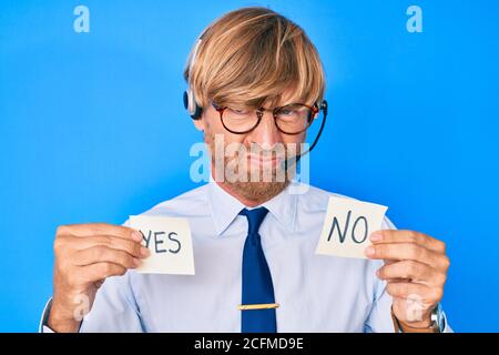 Giovane biondo uomo che indossa l'auricolare operatore che tiene sì e nessun promemoria scettico e nervoso, rondante sconvolto a causa del problema. Persona negativa. Foto Stock