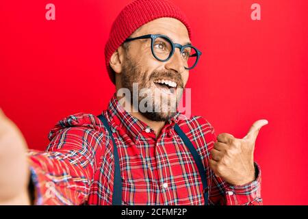 Bell'uomo hipster con barba scattando una foto selfie con smartphone che punta il pollice verso il lato sorridendo felice con aprire la bocca Foto Stock