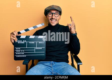 Bell'uomo con tatuaggi che tengono la clapboard del video film seduto sulla sedia del regista sorridendo con un'idea o domanda che punta il dito con il volto felice, num Foto Stock