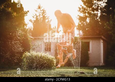 Padre e bambino saltano sopra l'acqua dall'irrigatore in giardino Foto Stock