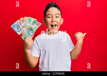 Bambino ispanico ragazzo piccolo che tiene i dollari australiani che punta il pollice in su al lato sorridente felice con bocca aperta Foto Stock