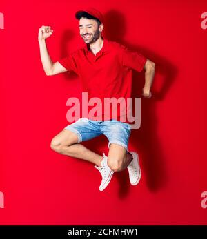 Giovane bel liberyman con cappuccio sorridente felice. Saltando con sorriso sul viso su sfondo rosso isolato Foto Stock