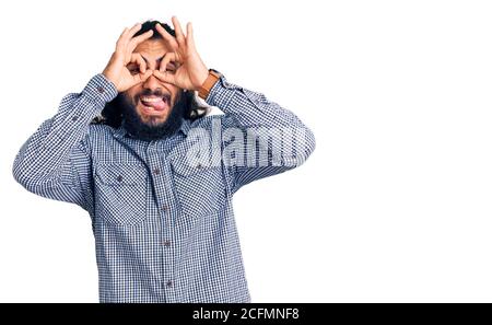 Giovane arabo che indossa abiti casual facendo un gesto ok come binocolo che si stacca la lingua, occhi che guardano attraverso le dita. Espressione pazzesca. Foto Stock