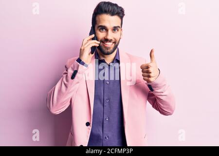 Giovane bell'uomo con barba che parla sullo smartphone sorridendo felice e positivo, il pollice in su facendo eccellente e segno di approvazione Foto Stock