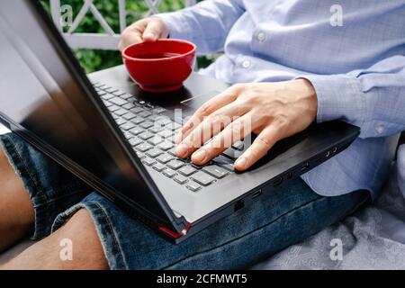 Immagine ritagliata di un giovane uomo che lavora sul suo computer portatile e beve caffè, vista posteriore delle mani di un uomo d'affari occupato utilizzando il computer portatile a casa. Foto Stock
