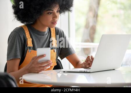 Giovane studentessa africana che usa un computer portatile che tiene uno smartphone seduto al tavolo. Foto Stock