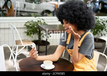 Donna africana felice che parla facendo la videochiamata al telefono al tavolo esterno del caffè. Foto Stock