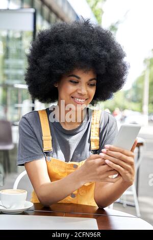 Sorridente donna hipster africana che usa il cellulare seduto al tavolo da caffè all'aperto. Foto Stock