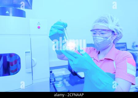 Una donna asiatica che indossa una maschera chirurgica che tiene un matraccio volumetrico o una provetta che fa la ricerca in un laboratorio. Messa a fuoco morbida su fl. Volumetrica Foto Stock