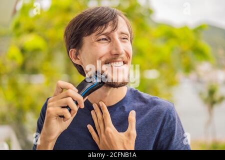 l'uomo sta radendo la barba con un rasoio elettrico sul balcone durante la quarantena. Bell'uomo bearded che rifilava la barba con un rifinitore a casa Foto Stock