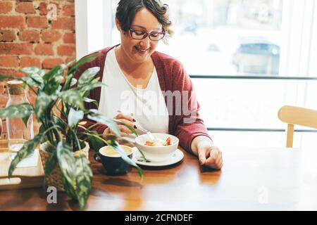 Donna bruna sorridente adulto in occhiali in abbigliamento casual Plus corpo di dimensioni positive con pranzo al caffè Foto Stock