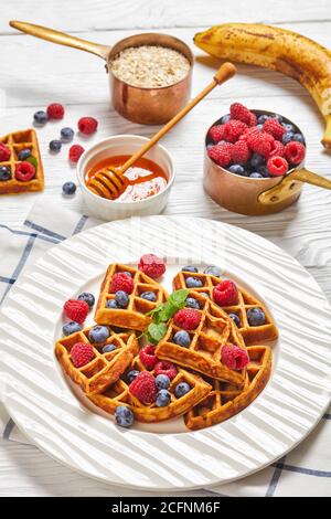 waffle alla banana d'avena serviti con frutti di bosco e miele su un piatto bianco su un tavolo di legno con ingredienti, frutti di bosco e miele, vista verticale dall'alto, Foto Stock