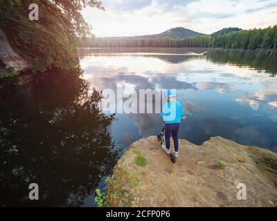 Sport blu che indossa ragazzo stare con divertimento spingere scooter sulla scogliera al lago e fa trucchi. Il sole serale all'orizzonte fa dei flares. Foto Stock