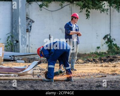 Mosca. Russia. 06 settembre 2020 i lavoratori in uniforme blu e caschi di sicurezza rossi in un cantiere stanno tagliando metallo con un macinacaffè. Scintille di Foto Stock