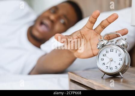 Stressato uomo africano che spegne sveglia sdraiato in letto all'interno Foto Stock