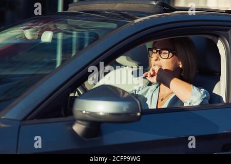 Felice donna matura che indossa occhiali tenendo in mano la carta di plastica vuota mentre guida la sua moderna auto attraverso la città in giornata di sole, guardando sul Foto Stock