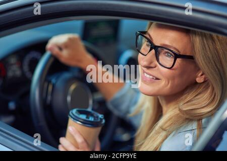 Sorridente uomo d'affari caucasico che indossa occhiali seduti dietro il volante di la sua auto moderna e l'acquisto take away caffè in carta tazza Foto Stock