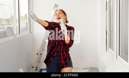 Felice giovane donna con camicia a scacchi rossa canta con pennello come microfono su una moderna e spaziosa terrazza a casa riparazione Foto Stock