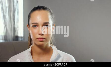 una giovane donna sportiva seria in felpa bianca guarda la fotocamera in primo piano della camera a casa Foto Stock