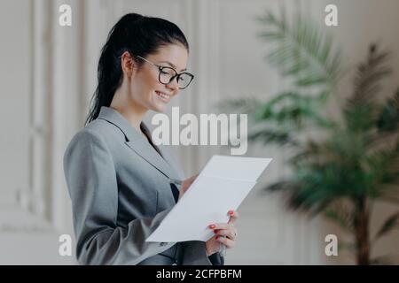 Felice avvocato donna scrive in documenti corporativi di carta, indossa occhiali ottici, abiti formali, stand in proprio armadio, sviluppa nuova strategia, ha felice Foto Stock