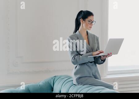 Foto di brunette executive lavoratore posa in casa ufficio vicino divano, tiene computer portatile, fa rapporto finanziario, indossa occhiali ottici per il decor della visione Foto Stock