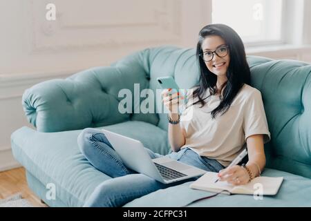 Felice studentessa lavora su carta corso, circondata da gadget moderni, fa note in blocco note, si sente rilassato a comodo divano, funziona online, wea Foto Stock