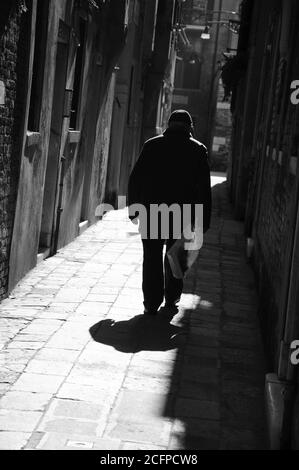 Silhouette di vecchio uomo che va su strada stretta in raggi di sole splendenti. (Venezia, Italia) un gioco di luce e ombra. Punti luce. Foto in bianco e nero. Foto Stock