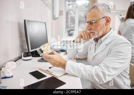 Dentista anziano esperto che tiene protesi dentali nel suo ufficio Foto Stock