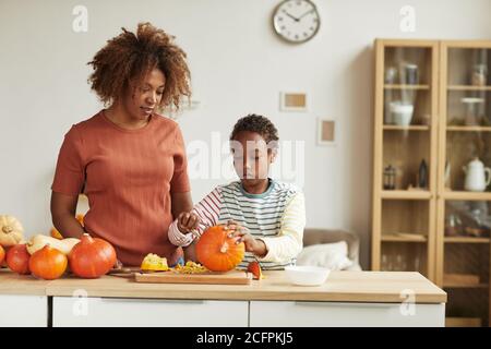 Giovane donna afroamericana adulta che indossa abiti casual in piedi a. tavola guardando il suo bambino che intagliano la zucca Foto Stock