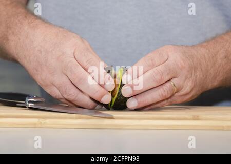 Primo piano delle mani maschili che separano le due metà di un avocado appena tagliato su un tagliere di legno Foto Stock