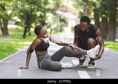 Caring Black Man massaggiando la gamba ferita della ragazza dopo la corsa Insieme all'aperto Foto Stock