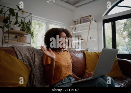 Sorridente giovane afroamericana che si rilassa sul divano agitando mentre parli con gli amici durante una videochiamata all'indirizzo home - giovane blogger che lavora da Foto Stock