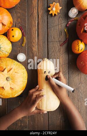 Immagine verticale dall'alto verso il basso di una persona irriconoscibile che disegna una faccia spaventosa Zucca con pennarello per Halloween Foto Stock