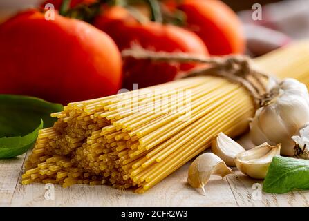 Spicchio di spaghetti crudi legato in un fascio con pomodori freschi di vite, aglio e basilico su un rustico tavolo di legno Foto Stock