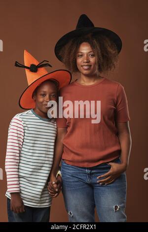 Verticale medio lungo studio ritratto di giovane donna moderna e. il suo figlio carino che indossa cappelli magici in piedi contro la parete marrone sfondo Foto Stock