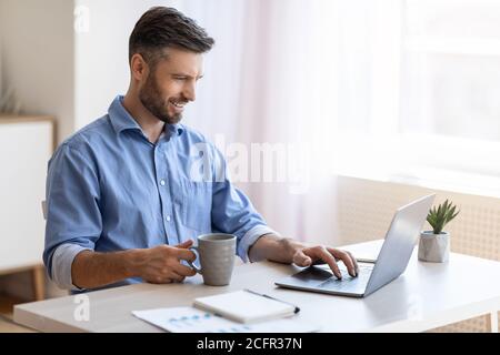 Ufficio domestico. Sorridente uomo freelancer controllare le e-mail sul computer portatile e bere caffè Foto Stock